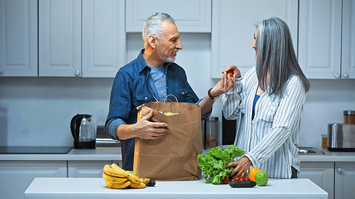 senior man giving cherry tomatoes to asian wife while unpacking paper bag in kitchen