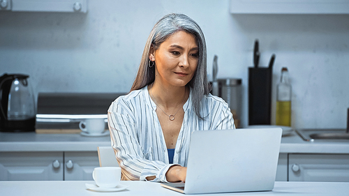 senior asian freelancer typing on laptop near coffee cup in kitchen