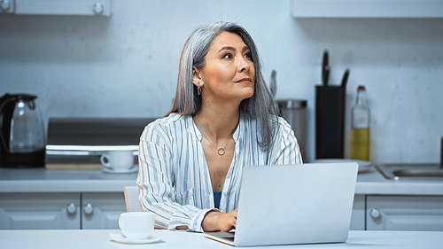 thoughtful asian freelancer working at laptop near cup of coffee in kitchen