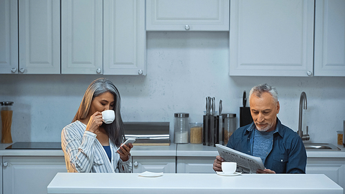 grey haired asian woman messaging on smartphone and drinking coffee near husband reading newspaper