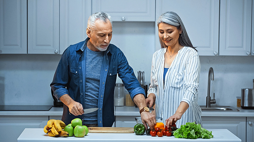 smiling interracial couple preparing breakfast with fresh vegetables and fruits in kitchen