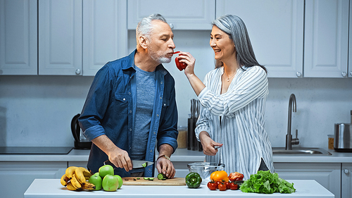 happy asian woman feeding husband with bell pepper while preparing breakfast in kitchen