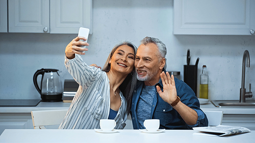cheerful elderly man waving hand near asian wife taking selfie in kitchen