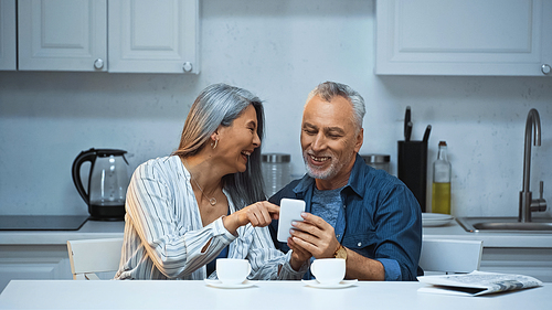 laughing asian woman pointing with finger at smartphone near smiling husband