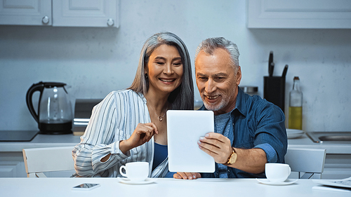 cheerful elderly interracial couple using digital tablet in kitchen