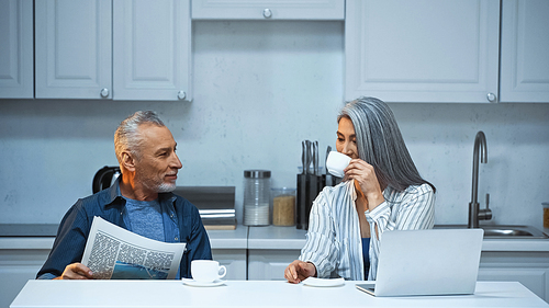 grey haired asian woman drinking coffee near husband with newspaper in kitchen