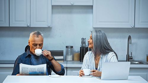smiling asian woman talking to elderly husband drinking coffee and reading newspaper in kitchen