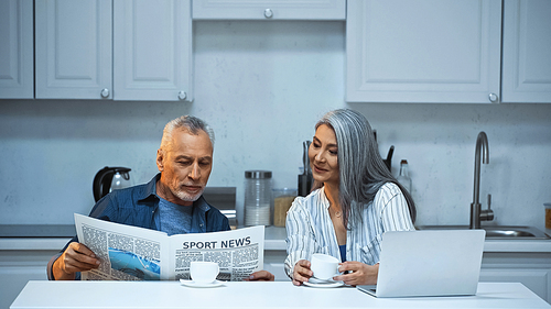smiling asian woman looking at elderly husband reading newspaper in kitchen