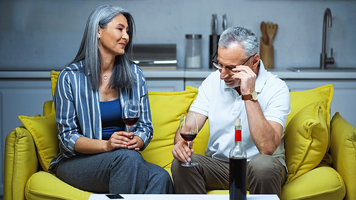 happy elderly interracial couple holding glasses with red wine on sofa at home