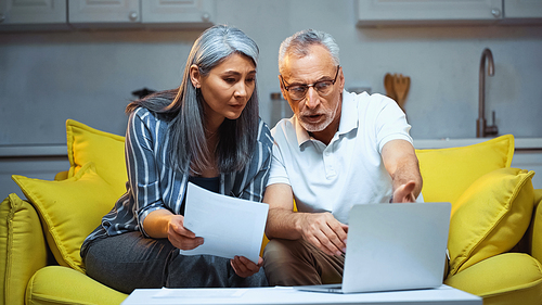 elderly asian woman holding document near husband pointing at laptop