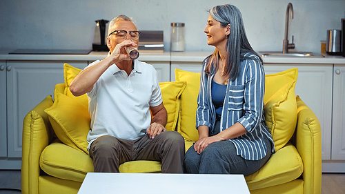elderly man drinking water near smiling asian wife sitting on couch at home