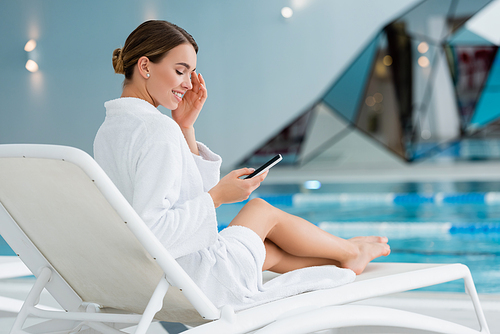 happy woman resting on deck chair and holding smartphone near pool