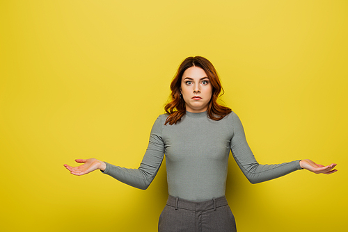 confused young woman with curly hair showing shrug gesture on yellow