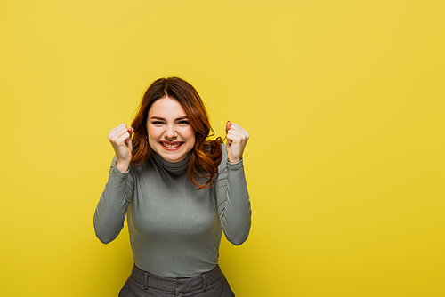 excited woman with curly hair standing with clenched fists isolated on yellow