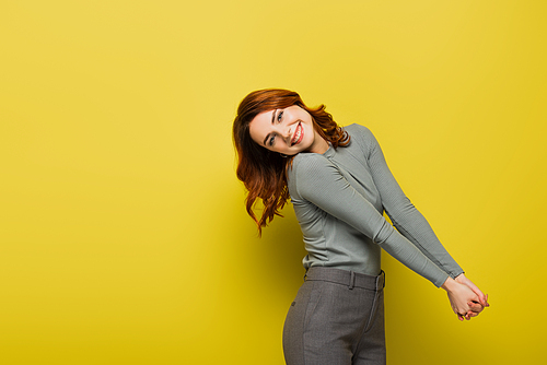 cheerful woman with curly hair standing with clenched hands on yellow