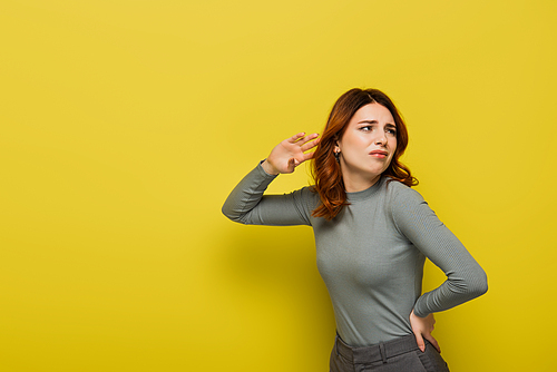 displeased woman with curly hair standing with hand on hip while listening on yellow