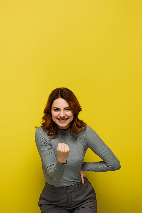 curly woman with clenched fist and hand on hip standing and smiling on yellow