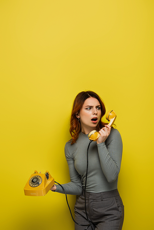 shocked young woman holding retro telephone and looking away isolated on yellow