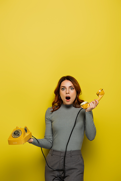 shocked young woman holding retro telephone isolated on yellow