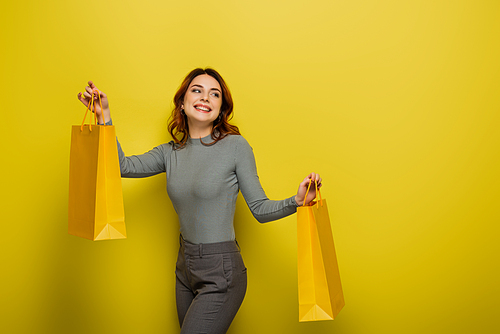 happy young woman with curly hair holding shopping bags on yellow