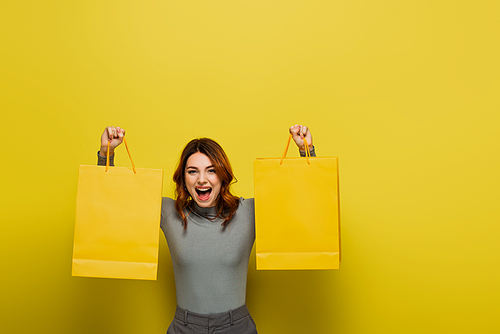 amazed young woman with curly hair holding shopping bags on yellow