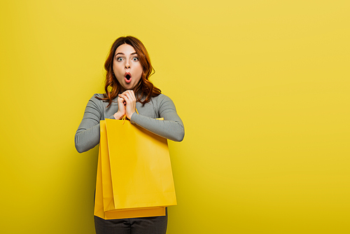 shocked young woman with curly hair holding shopping bags on yellow