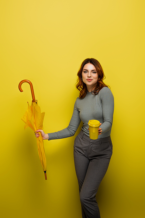 happy young woman with curly hair holding paper cup and umbrella on yellow