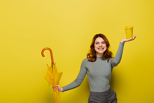 happy woman with curly hair holding paper cup and umbrella on yellow