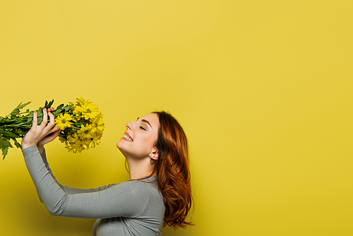 cheerful young woman with wavy hair holding flowers on yellow