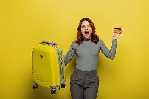 amazed woman with curly hair holding luggage and credit card on yellow