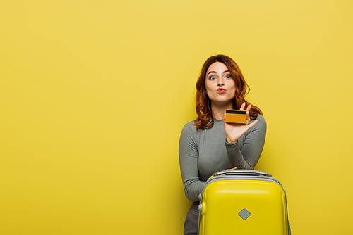 woman with curly hair pouting lips while holding baggage and credit card on yellow
