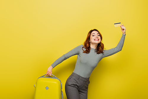 cheerful woman with curly hair holding baggage and credit card on yellow