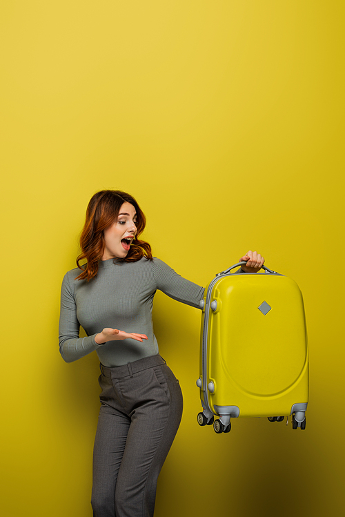 amazed woman with curly hair pointing with hand at luggage on yellow