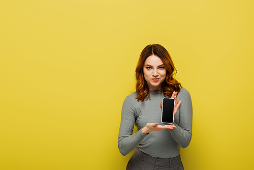 cheerful woman with curly hair holding smartphone with blank screen on yellow