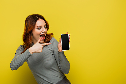 amazed woman with curly hair pointing with finger at smartphone with blank screen on yellow