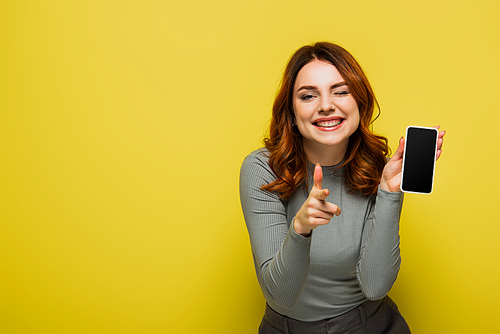 cheerful woman holding smartphone with blank screen and pointing with finger while  on yellow