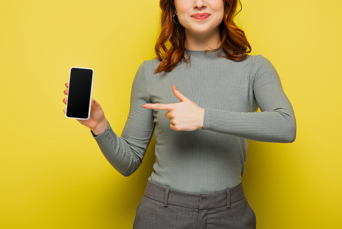 cropped view of cheerful woman with curly hair pointing with finger at smartphone with blank screen on yellow