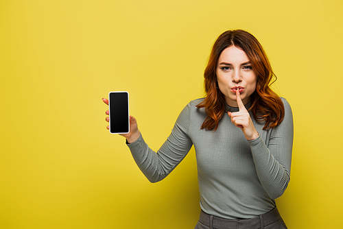 woman with curly hair showing hush sign and holding smartphone with blank screen on yellow