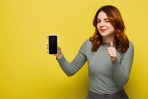 cheerful woman with curly hair holding smartphone with blank screen and showing thumb up on yellow