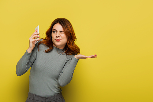 displeased young woman with curly hair winking eye while holding smartphone on yellow