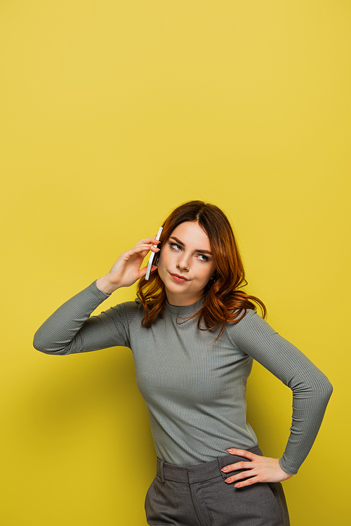 bored young woman with curly hair talking on smartphone and standing with hand on hip on yellow