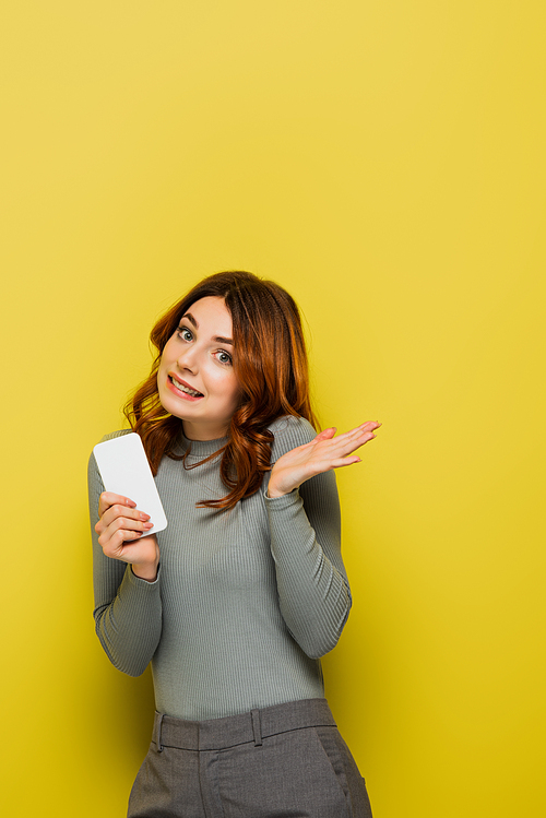 clueless young woman with curly hair holding smartphone and gesturing on yellow