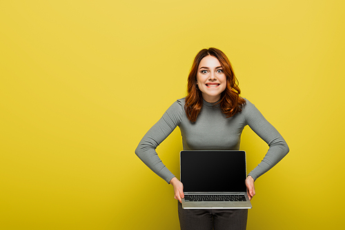cheerful young woman with curly hair holding laptop with blank screen and biting lips on yellow