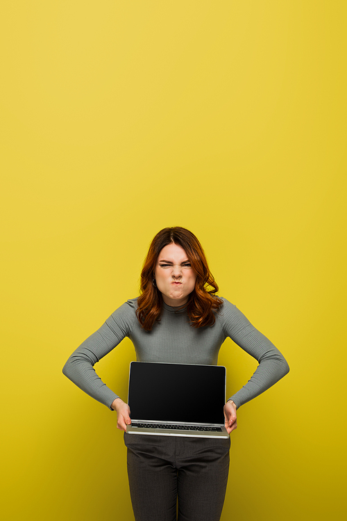 irritated young woman with curly hair holding laptop with blank screen on yellow