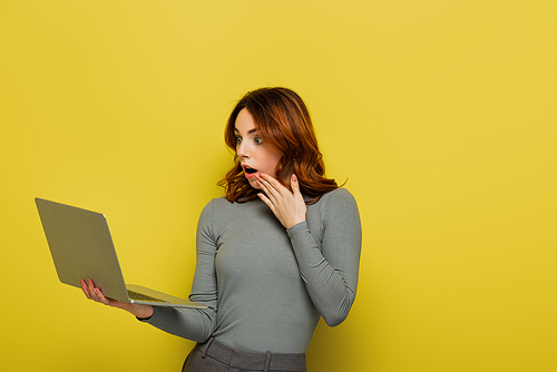 shocked young woman with curly hair holding laptop isolated on yellow