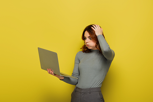 shocked woman with curly hair puffing cheeks while holding laptop on yellow