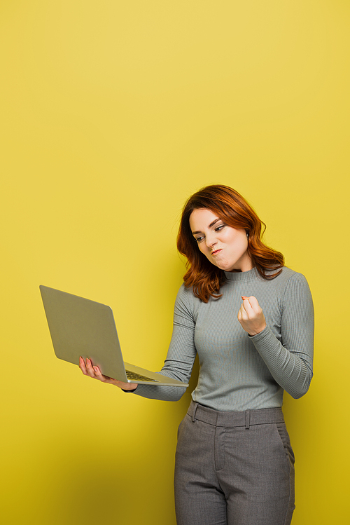 angry young woman with curly hair holding laptop and showing clenched fist on yellow