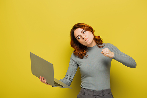 mad young woman with curly hair holding laptop and showing clenched fist on yellow