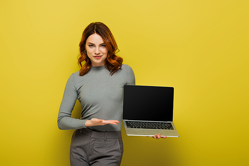 young woman with curly hair pointing with hand at laptop with blank screen on yellow