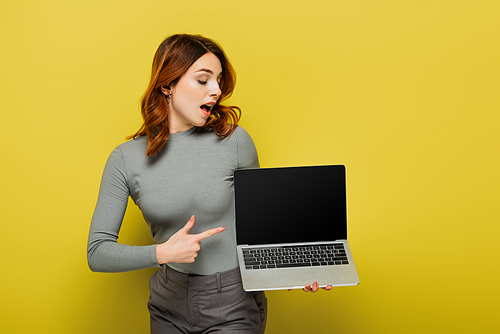 amazed woman with curly hair pointing with finger at laptop with blank screen on yellow